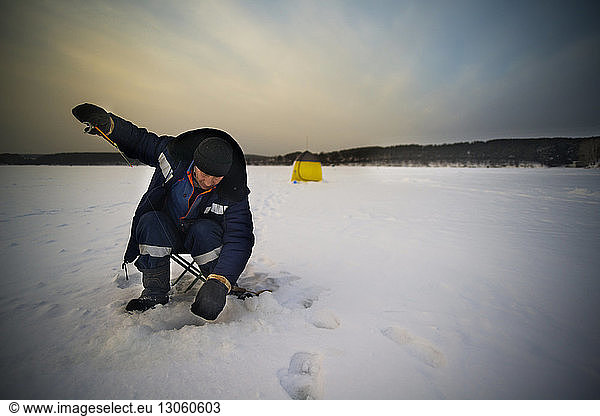 Mature male ice fishing on frozen lake against sky at sunset