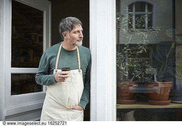 Mature male owner looking away while having coffee at entrance of art studio