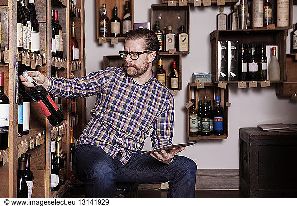 Mature male small business owner reading label on wine bottle while holding tablet computer in shop