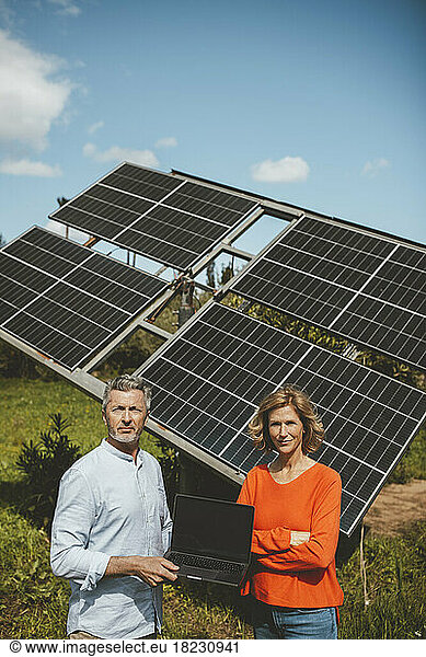 Mature man and woman standing with laptop in front of solar panels on sunny day