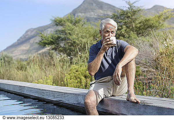 Mature man drinking coffee while sitting by swimming pool against mountain