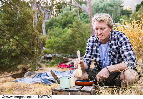 Mature man frying breakfast on camping stove in forest, Deer Park, Cape Town, South Africa