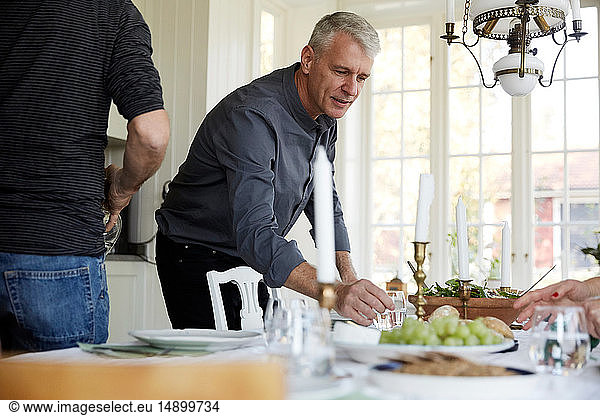 Mature man holding drinking glass on dining table by friend at home