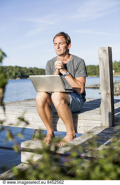 Mature man praying while sitting with laptop on pier