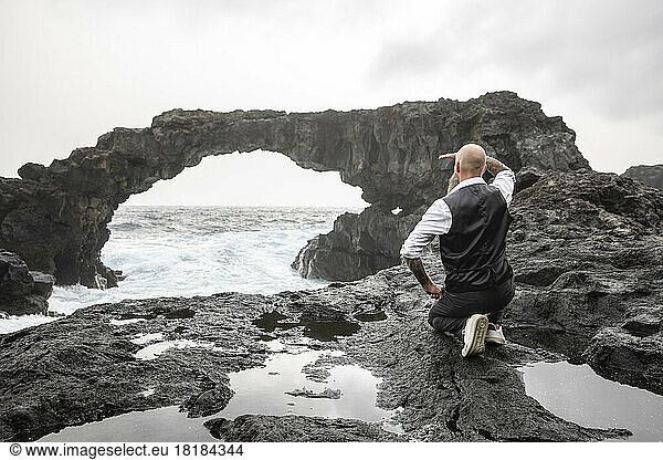 Mature man shielding eyes kneeling in front of rock arch