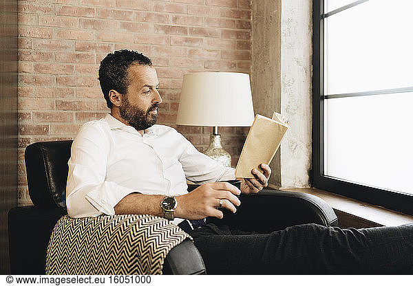 Mature man sitting in armchair by window  reading book