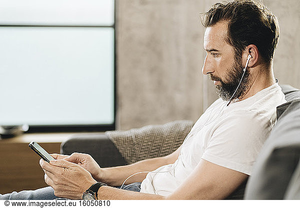 Mature man sitting on couch  using smartphone and earphones