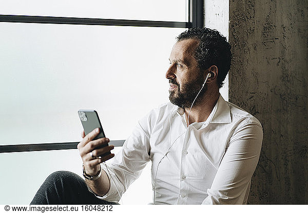 Mature man sitting on window sill  using smartphone and earphones