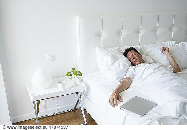 Mature man sleeping by laptop in bed