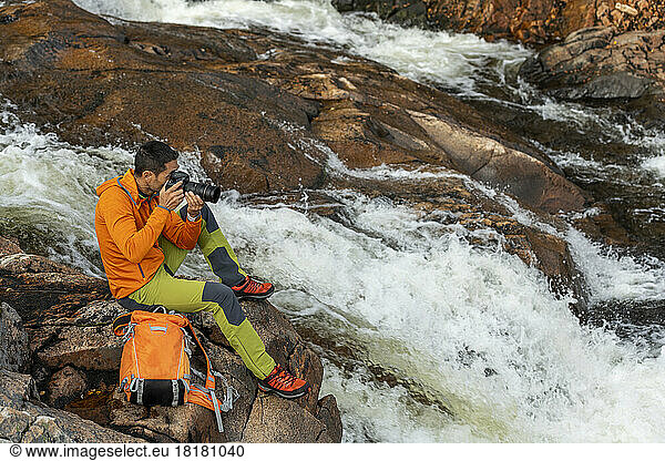 Mature man taking photos through camera sitting on rock by river