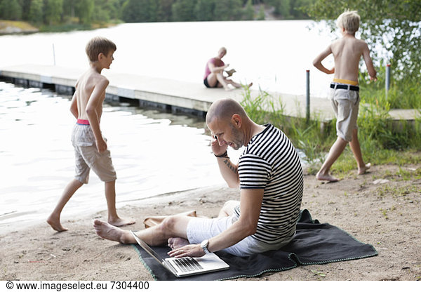 Mature man using laptop while communicating on cell phone at beach with family in the background
