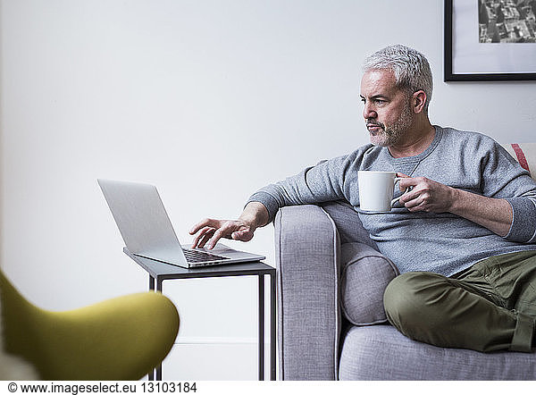 Mature man using laptop while holding coffee mug at home