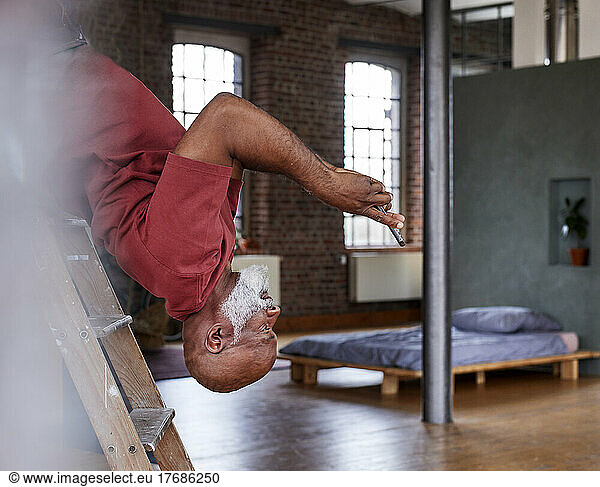 Mature man using smart phone and bending backwards on ladder in bedroom