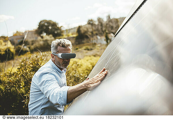 Mature man wearing VR glasses touching solar panel in garden