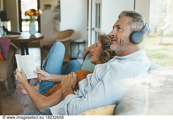 Mature man wearing wireless headphones sitting by woman with book