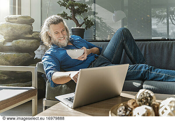 Mature man with a beard eating a salad and looking at his laptop on a