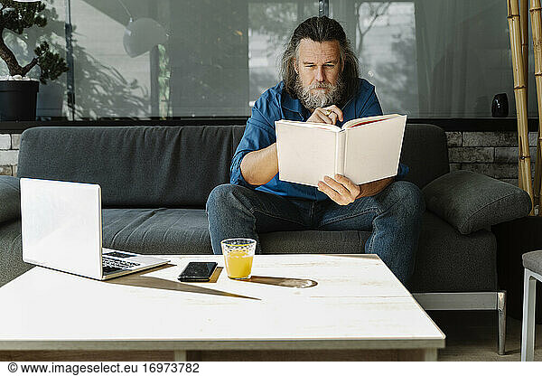 Mature man with a beard reading a book sitting on a sofa in the living