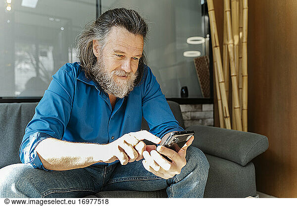 Mature man with a beard sitting on a sofa and looking at his smartphone in the living room of his home. Business concept