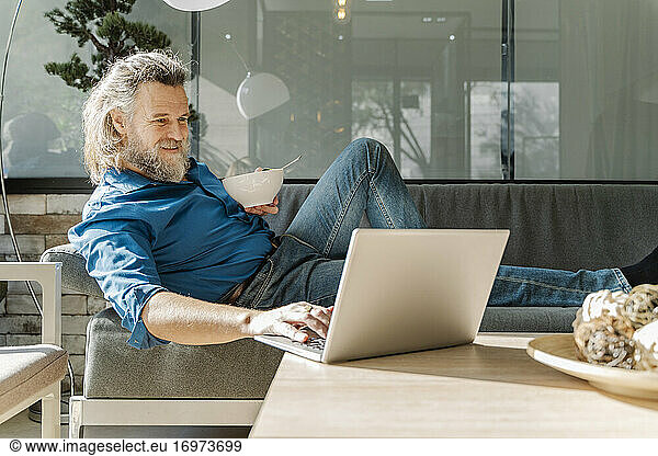 Mature man with a beard smiling and working with his laptop on a sofa