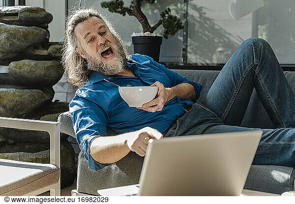 Mature man with a beard smiling and working with his laptop on a sofa