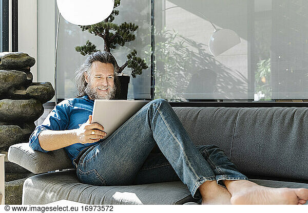 Mature man with a beard smiling and working with his laptop on a sofa at home. Business concept
