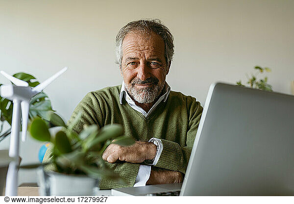 Mature man with arms crossed using laptop while sitting by table at home