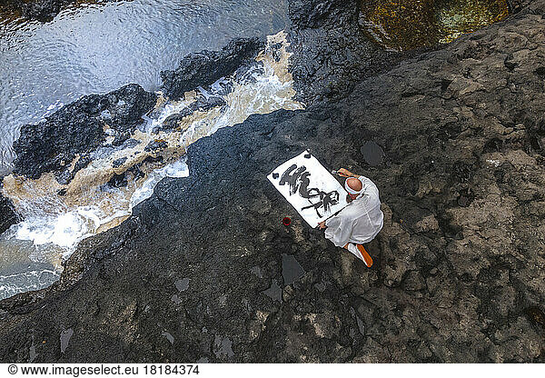 Mature monk doing calligraphy sitting on rock by sea