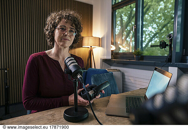 Mature presenter wearing eyeglasses holding headset sitting with laptop at desk in radio station