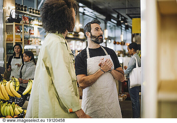 Mature sales clerk with hands clasped standing by male customer at grocery store