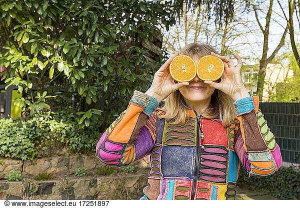 Mature woman covering eyes with oranges halved in hand outdoors