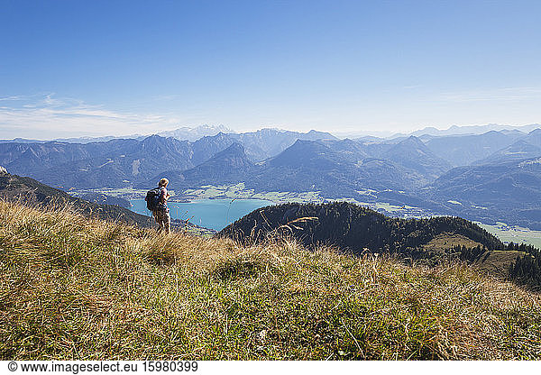 Mature woman hiking on grass at Schafberg Railway with Lake Wolfgangsee and Dachstein Mountains against blue sky