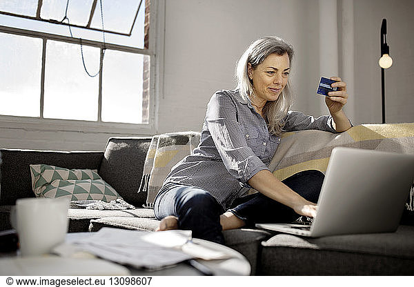 Mature woman holding debit card while using laptop on sofa