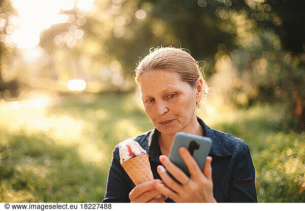 Mature woman holding smart phone and ice cream standing in park