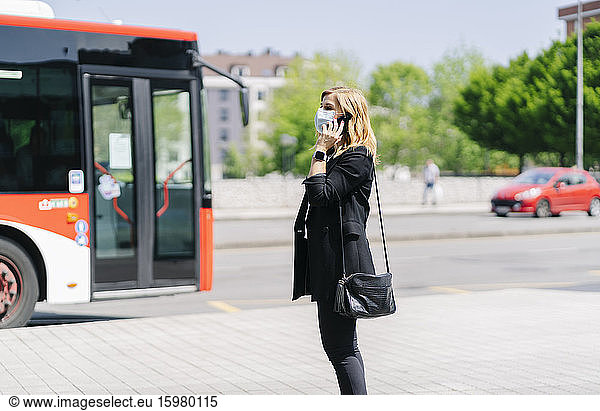 Mature woman on the phone wearing protective mask waiting at bus stop  Spain