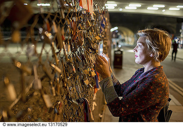 Mature woman photographing love locks with digital tablet at night  London  UK
