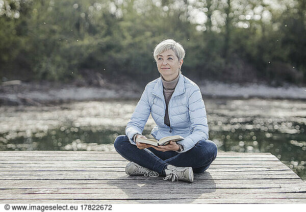 Mature woman sitting cross-legged with book on pier