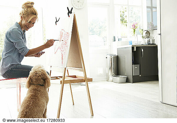 Mature woman sitting with dog while painting on canvas at home