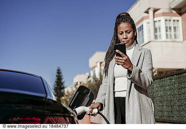 Mature woman using smart phone and charging car on sunny day