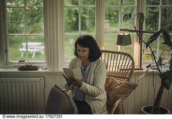 Mature woman using smart phone while sitting on chair near window at home