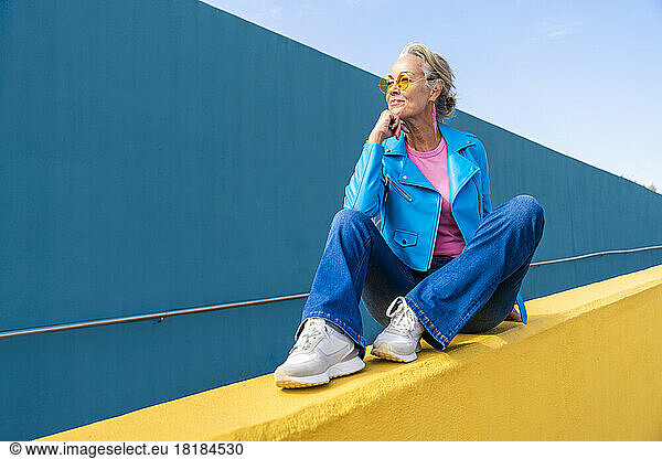 Mature woman wearing blue leather jacket sitting on yellow wall