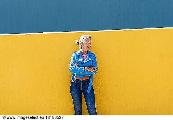 Mature woman with arms crossed leaning on colored wall