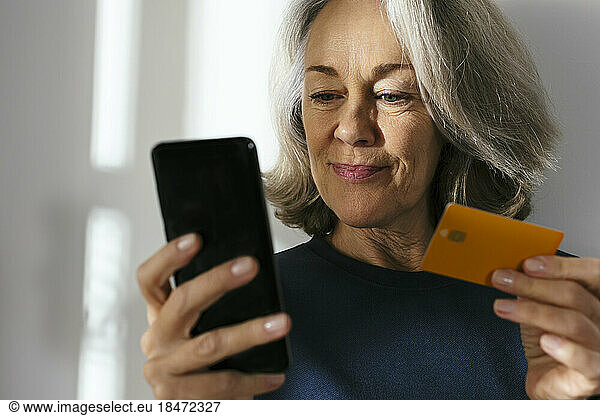 Mature woman with credit card using smart phone in front of wall