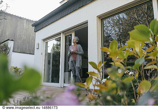 Mature woman with cup of coffee standing at terrace door