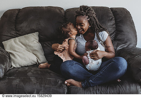 Mehrrassige 2 Jahre alte  küssende Mutter mit Zöpfen  die ihr Neugeborenes auf der Couch hält