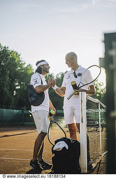 Men exchanging handshake holding tennis racket while standing at sports court