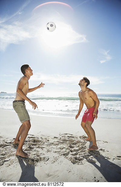 Men in swim trunks heading soccer ball on beach