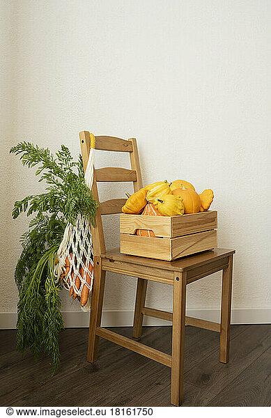 Mesh bag with carrots and crate of pumpkins on chair in front of wall