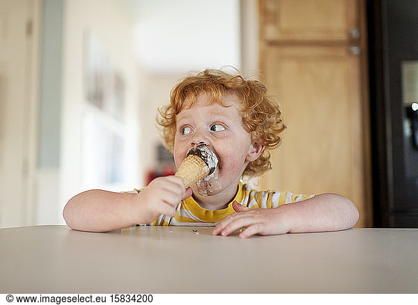 Messy toddler boy eats ice cream while sitting at counter in kitchen