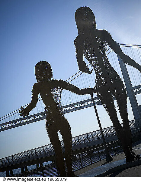 Metal sculpture of two people with a bridge in the background.