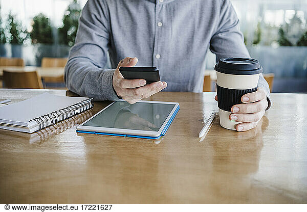 Mid adult businessman with reusable coffee cup using smart phone on desk at office